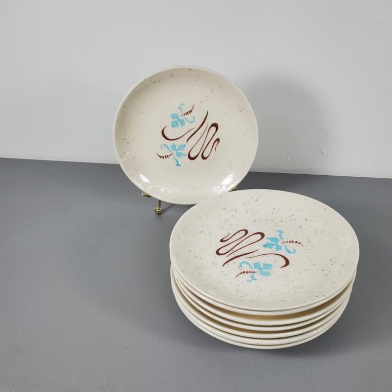May include: A stack of eight white ceramic dessert plates with a speckled glaze. The plates have a brown and blue floral design with a brown swirl.