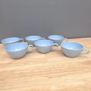 May include: Six light blue teacups with handles are arranged on a wooden surface. The cups have a rounded shape with a slightly angled rim. The interior of the cups is a lighter shade of blue than the exterior.
