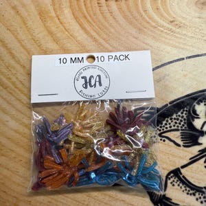 May include: A clear plastic bag containing a 10 pack of 10 mm fishing lures in various colors including purple, gold, red, and blue, all featuring a glitter finish. The header card displays the text 10 MM 10 PACK and a logo for JCA Fishing Lures.