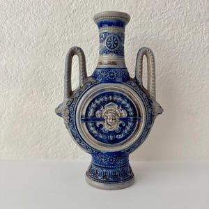 Puede incluir: Una vasija de cerámica azul y gris con dos asas laterales, un cuello estrecho y un medallón central circular. El medallón muestra un relieve de un rostro humano rodeado de volutas ornamentadas y motivos florales en esmalte azul cobalto.