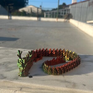 May include: A flexible, articulated dragon figurine featuring a metallic, iridescent finish that shifts between vibrant green, gold, and deep red hues. The creature has a detailed head with horns and a long, segmented body with raised scales.