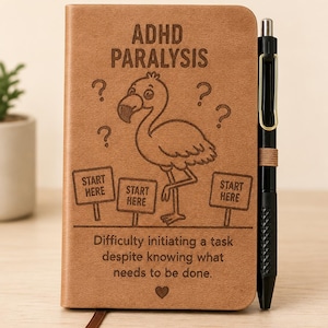 May include: A tan notebook with a black pen attached to the side. The cover features an engraved flamingo, question marks, three signs reading START HERE, and the text ADHD PARALYSIS and Difficulty initiating a task despite knowing what needs to be done.