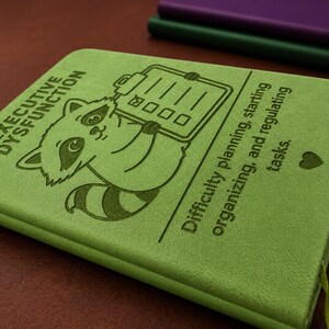 Peut inclure: Un carnet vert clair avec une illustration grav&eacute;e en vert fonc&eacute; repr&eacute;sentant un raton laveur tenant un presse-papier. Le texte indique : EXECUTIVE DYSFUNCTION. Difficulty planning, starting, organizing, and regulating tasks. Un petit c&oelig;ur.