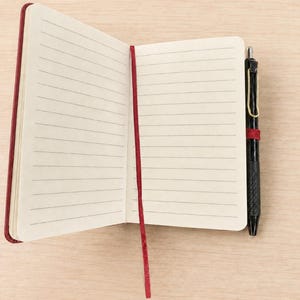 Peut inclure: Un carnet ouvert avec des pages lign&eacute;es de couleur cr&egrave;me et un marque-page en ruban rouge fonc&eacute;. Un stylo r&eacute;tractable noir avec un clip dor&eacute; est maintenu dans une boucle &eacute;lastique rouge fonc&eacute; assortie fix&eacute;e sur le c&ocirc;t&eacute; de la couverture.