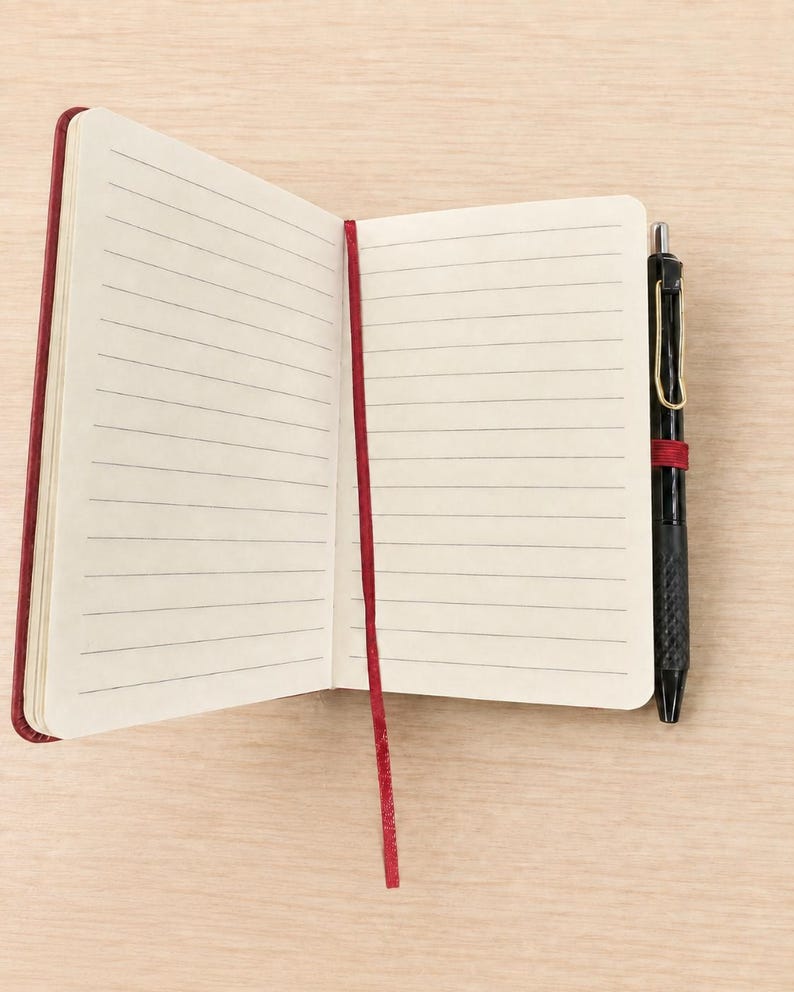 Peut inclure: Un carnet ouvert avec des pages lign&eacute;es de couleur cr&egrave;me et un marque-page en ruban rouge fonc&eacute;. Un stylo r&eacute;tractable noir avec un clip dor&eacute; est maintenu dans une boucle &eacute;lastique rouge fonc&eacute; assortie fix&eacute;e sur le c&ocirc;t&eacute; de la couverture.