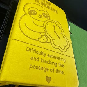 May include: A bright yellow notebook with a textured cover featuring an engraved illustration of a sloth holding a melting clock. The text reads TIME BLINDNESS and Difficulty estimating and tracking the passage of time, with a small heart icon below.