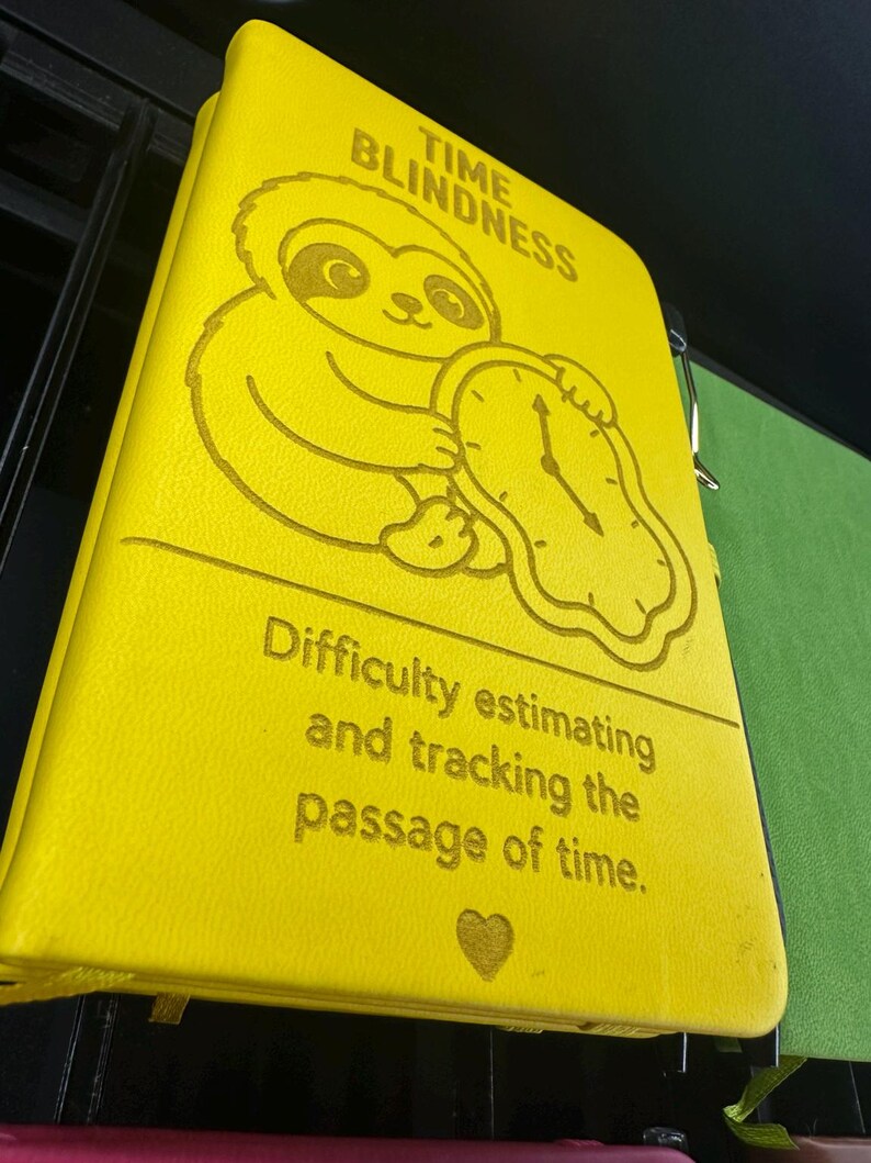 May include: A bright yellow notebook with a textured cover featuring an engraved illustration of a sloth holding a melting clock. The text reads TIME BLINDNESS and Difficulty estimating and tracking the passage of time, with a small heart icon below.