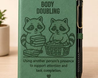 Peut inclure: Un carnet vert avec un stylo noir attaché sur le côté. La couverture présente deux ratons laveurs illustrés, le texte "BODY DOUBLING" et "Using another person's presence to support attention and task completion," ainsi qu'un petit cœur.