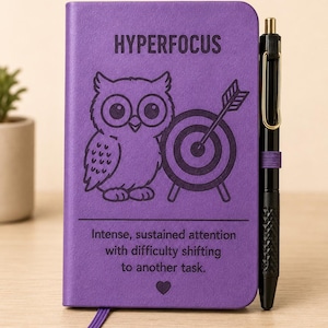 May include: A purple notebook with a black pen attached to the side. The cover features an owl illustration, a target, and the text "HYPERFOCUS" above "Intense, sustained attention with difficulty shifting to another task." with a small heart icon.
