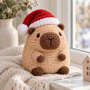 May include: A crochet capybara plush toy featuring a tan body, dark brown paws, ears, and snout, with small black eyes and pink cheeks. The capybara wears a red and white crocheted Santa hat with a white pom-pom on top, crafted from soft yarn.