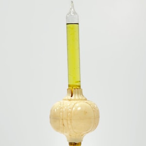 May include: A vertical glass tube filled with a translucent yellow liquid, set into a cream-colored, spherical base with a ribbed, segmented texture. The glass tube is sealed at the top with a pointed tip, and the base features a decorative, rounded design.