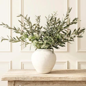 May include: A white ceramic vase with a textured, matte finish containing a large arrangement of green olive branches and small clusters of white flowers. The vase has a rounded, bulbous shape and sits upright, displaying a natural botanical display.
