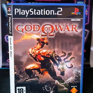 Może przedstawiać: Niebieskie plastikowe pudełko na grę wideo PlayStation 2 God of War. Okładka zawiera tytuł napisany czerwoną i białą czcionką, postać trzymającą świecącą głowę Meduzy, logo PlayStation 2 oraz symbol ograniczenia wiekowego 18+ w rogu.