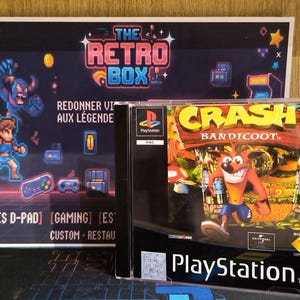 Può includere: Una custodia per videogioco PlayStation di Crash Bandicoot con il personaggio arancione sulla copertina, posizionata davanti a un cartello rettangolare con il testo The Retro Box e varie illustrazioni in pixel art di personaggi.