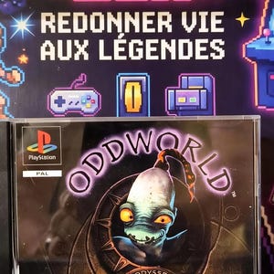 Puede incluir: Una caja de videojuego de PlayStation para Oddworld: Lodysee d'Abe que presenta un personaje central con grandes ojos naranjas y una cabeza pálida. La portada incluye el logotipo de PlayStation, el título y una pegatina de The Retro Box.
