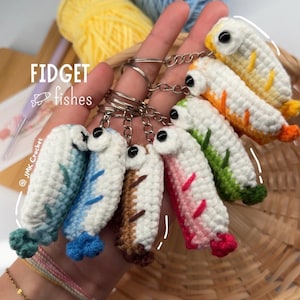 May include: A collection of seven small, handmade crochet fish keychains, each featuring a white body with colorful accents in yellow, orange, green, pink, brown, blue, and teal. The text FIDGET fishes and JMK Crochet is printed on the image.