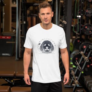 May include: A light grey short-sleeved t-shirt featuring a black ink-style graphic of a dog face surrounded by small stars and a tiny planet. The design is centred on the chest, showcasing a detailed illustration of a canine with a happy expression.
