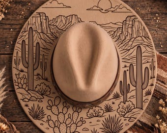 May include: A tan felt hat featuring a dark brown leather band with metallic accents. The wide brim is decorated with a black line art illustration of a desert landscape, including saguaro cacti, prickly pear cacti, and distant mountains.