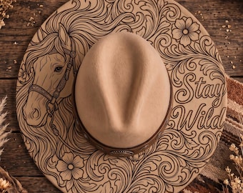 May include: A beige felt cowboy hat featuring a dark brown leather band with metallic accents. The wide brim is decorated with a detailed dark brown laser-engraved design of a horse head, floral scrollwork, and the text Stay Wild in script.