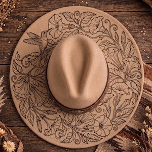 May include: A beige wide-brimmed hat featuring a dark brown leather band and intricate black line art illustrations on the brim. The design depicts a hummingbird hovering near large trumpet-shaped flowers surrounded by swirling leafy vines.