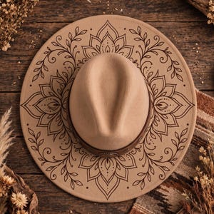 Puede incluir: Un sombrero de ala ancha en color beige con un diseño de mandala impreso en marrón oscuro sobre el ala. El sombrero tiene una copa central con un pliegue y una banda de aspecto de cuero marrón oscuro alrededor de la base, decorada con pequeños remaches metálicos.