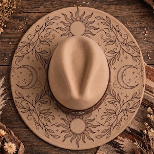 May include: A beige wide-brimmed hat featuring dark brown line art of suns, crescent moons, stars, and leafy vine patterns on the brim. A brown leather band with metallic accents encircles the base of the crown, which has a central crease.