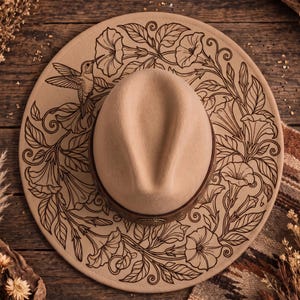 Puede incluir: Un sombrero beige de ala ancha con una banda de cuero marrón oscuro e ilustraciones de líneas negras en el ala. El diseño muestra un colibrí volando cerca de grandes flores en forma de trompeta rodeadas de enredaderas con hojas.