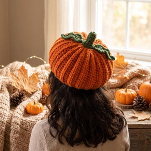 Pode incluir: Um gorro de crochê em forma de abóbora na cor laranja vibrante, com um design texturizado canelado e um caule de crochê verde-escuro com duas folhas verdes presas no topo. O gorro é feito com fio grosso e tem uma aparência arredondada e macia.