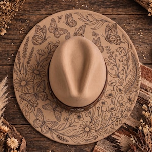 Puede incluir: Un sombrero beige de ala ancha con una banda de cuero marrón oscuro y un concho metálico central. El ala está decorada con intrincadas ilustraciones de líneas negras de varios tipos de mariposas, flores silvestres, margaritas y helechos.
