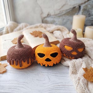 Puede incluir: Tres calabazas de ganchillo con tallos marrones. La calabaza de la izquierda es naranja con un patrón de goteo marrón. La central es naranja con ojos y nariz negros. La de la derecha es marrón con ojos y boca de color naranja brillante.