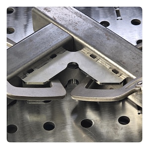 May include: A silver-coloured metal corner welding jig holding two pieces of square steel tubing at a ninety-degree angle. The jig features a flat L-shaped plate with integrated clamps, positioned on a perforated metal welding table surface.
