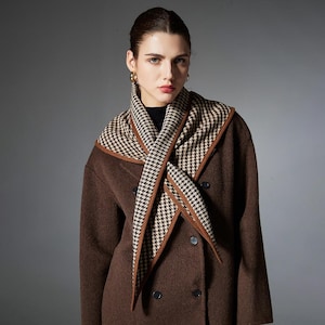 May include: A brown double-breasted coat with a dark interior layer and a triangular scarf featuring a black and beige houndstooth pattern. The scarf has a solid brown border and is draped around the neck, layered over the coat's wide lapels.