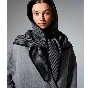May include: A hooded scarf featuring a black and grey diagonal striped pattern with a solid black trim. The scarf is draped over the head and shoulders, layered over a solid black top and a grey wool-blend coat with visible black buttons.