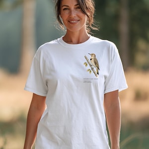 May include: A white short-sleeve t-shirt featuring a detailed illustration of a Northern Flicker bird perched on a branch. The text below the bird reads: NORTHERN FLICKER - FEMALE, Colaptes Auratus, NORTH AMERICA. The shirt has a crew neckline.