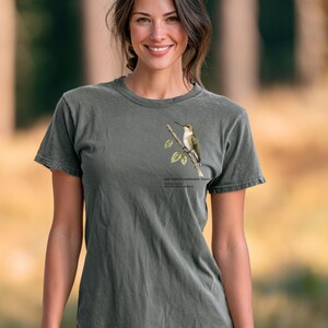 May include: A charcoal grey short-sleeve t-shirt featuring a graphic of a ruby-throated hummingbird perched on a branch with leaves. The text below the bird reads: RUBY-THROATED HUMMINGBIRD - FEMALE, ARCHILOCHUS COLUBRIS, EASTERN NORTH AMERICA.