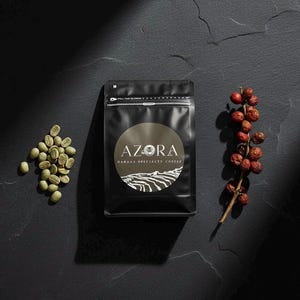 Op de afbeelding: Een zwarte koffiezak met een rond etiket met de tekst AZORA HAWAAJ SPECIALTY COFFEE en een afbeelding van terrasvormige velden. Links ligt een hoopje lichtgroene rauwe koffiebonen en rechts een takje met kleine rode koffiebessen.