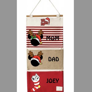 May include: A hanging fabric wall organiser with three pockets. The top pocket has red stripes, a reindeer-eared character, and the text MOM. The middle pocket is tan with a reindeer-eared character and DAD. The bottom is red with a puppy in a stocking and JOEY. The top features the text HO HO HO.
