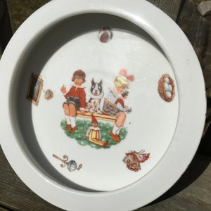 Op de afbeelding: Witte porseleinen kinderkom met een rand. De kom heeft een kleurrijke illustratie van drie kinderen die op een bankje zitten met een hond en een vogel. De illustratie bevat een verscheidenheid aan andere details, zoals een fotolijst, een nest met eieren, een vlinder en een sleutel.