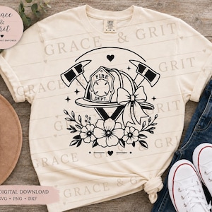May include: A cream-coloured short-sleeve t-shirt featuring a black graphic print of a firefighter helmet, two crossed axes, a bow, and floral accents. The helmet is labelled FIRE DEPT. The design includes small heart and star icons surrounding the central elements.