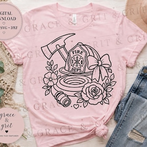 May include: A light pink t-shirt featuring a black line art graphic of a firefighter helmet with the text FIRE DEPT, a crossed axe, a coiled fire hose, a large bow, and floral accents. The shirt is displayed with blue denim jeans.
