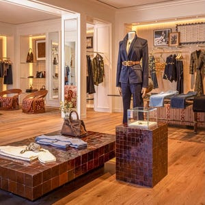 May include: A retail display featuring a dark navy blazer and matching trousers on a mannequin, accessorised with a tan leather belt. A brown leather handbag and folded garments rest on a low, square, dark brown tiled table near a glass display case.