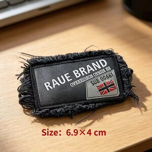 May include: A rectangular black fabric patch with frayed edges featuring the white text RAUE BRAND, OVER HOROS OYNIIE ES, and SUB.00661, along with a small Union Jack flag graphic. The patch measures 6.9 by 4 cm as indicated in the image.
