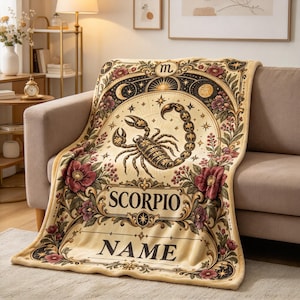 May include: A cream-colored throw blanket featuring a central black scorpion illustration, celestial motifs including moons and stars, and floral borders in shades of red and green. The text SCORPIO and NAME are printed in black serif font.
