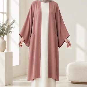 Peut inclure: Un cardigan ouvert de style kimono rose poudré avec des manches larges et des poches latérales, porté sur une robe sans manches crème arrivant au sol. L'ensemble est assorti d'un foulard rose poudré et de chaussures à bout pointu.