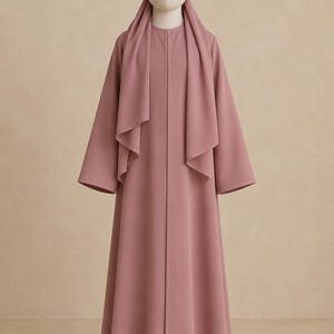 Peut inclure: Une abaya rose poudré dotée d'une encolure ronde, de manches longues et d'un foulard assorti drapé sur les épaules. Le vêtement présente une silhouette droite arrivant au sol avec une fermeture frontale nette et sans couture.