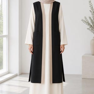 May include: A long, sleeveless black vest featuring beige trim along the front opening and two front pockets, worn over a floor-length cream-colored dress with long sleeves and a matching cream-colored headscarf covering the head and neck.