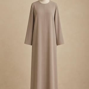 Op de afbeelding: Een vloerlange jurk met lange mouwen in een effen taupe kleur. Het kledingstuk heeft een eenvoudige ronde halslijn en een recht, strak silhouet dat tot op de grond valt, weergegeven op een neutraal gekleurde paspop.