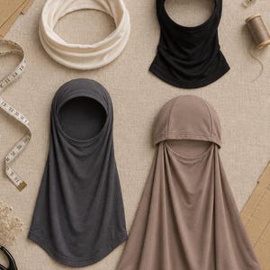 May include: A collection of head coverings including a cream-colored neck gaiter, a black hood, a dark gray hood, and a light brown hood. The items are arranged alongside sewing tools such as a measuring tape, scissors, and thread spools.