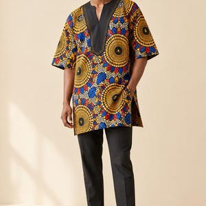 May include: A short-sleeved tunic featuring a vibrant, repeating circular pattern in yellow, blue, and red on a white background. The garment has a solid black V-neck collar and side slits at the hem, paired with plain black trousers and black loafers.