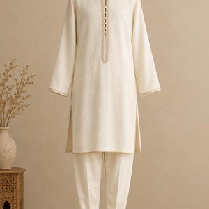 May include: A cream-colored long-sleeve tunic and matching trousers set. The tunic features a round neckline with a vertical placket containing five small buttons, side slits at the hem, and light beige trim along the cuffs and collar.